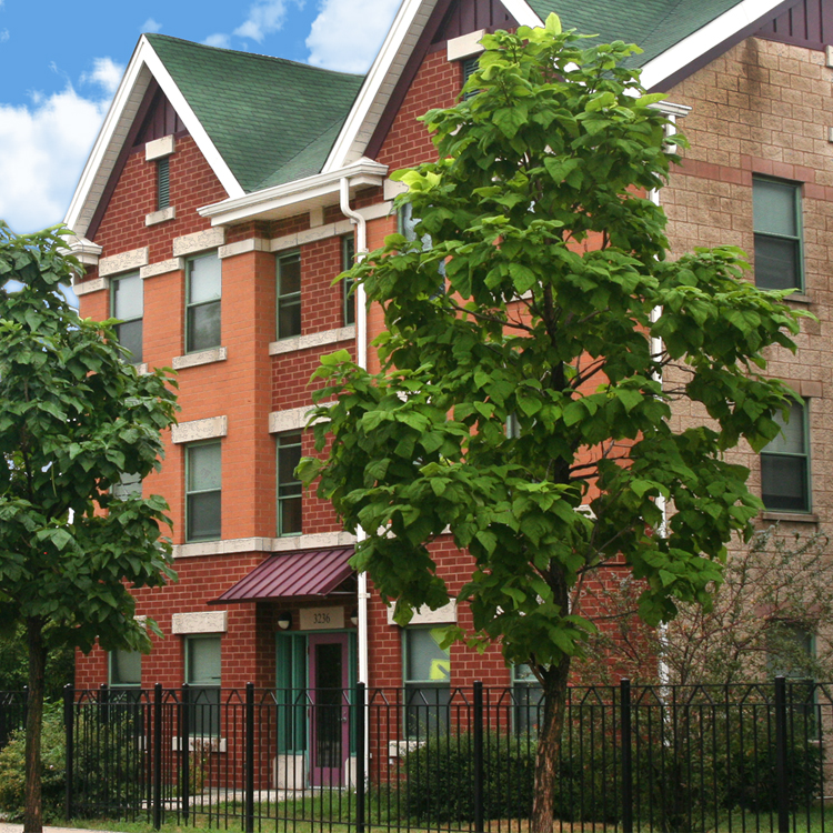 Three-story, red brick, highly energy efficient home built by Claretian Associates with a wrought iron fence and two trees in front. 