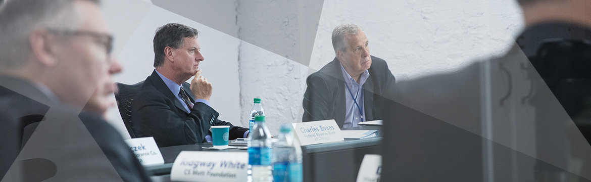 Charles Evans, among others, sits at a table, listening intently to someone out of frame. 